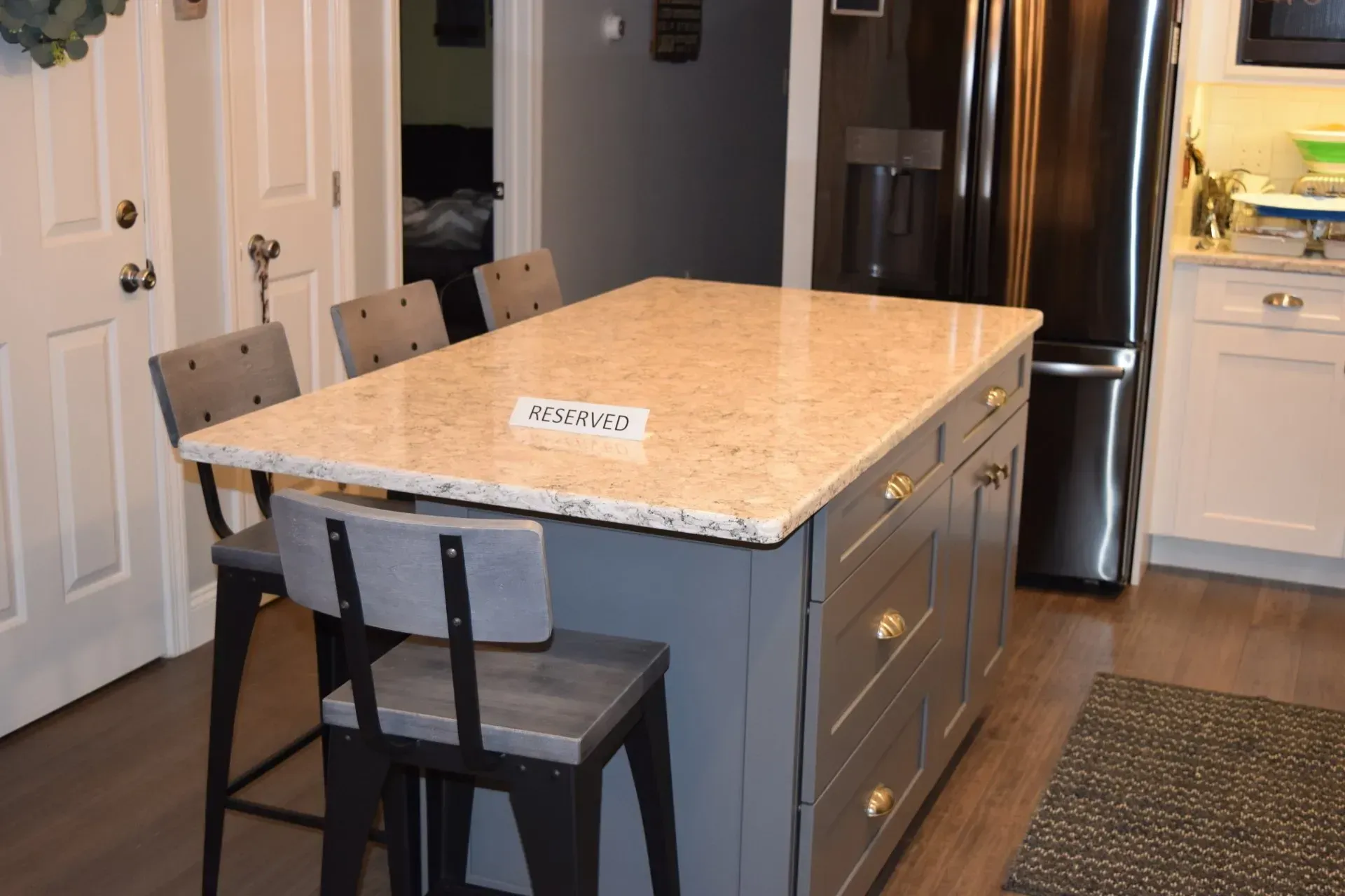 Kitchen island with granite countertop, gray cabinets, and three bar stools. Black refrigerator in the background.