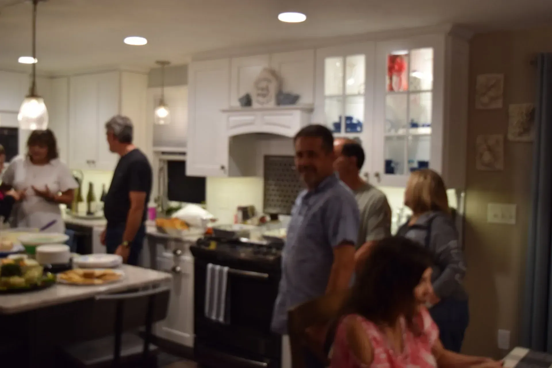 People gathered in a well-lit kitchen. Some are standing, others seated at a table. Food on counters.