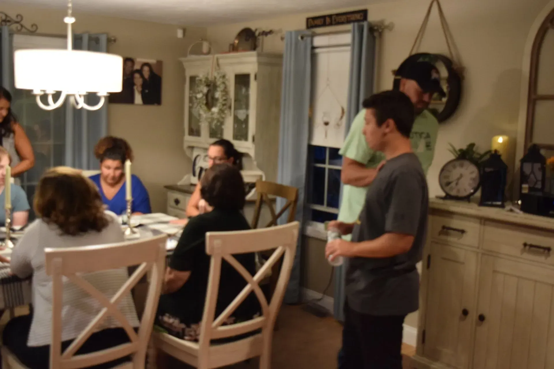 People gathered around a dining table in a warmly lit room. Others stand nearby.