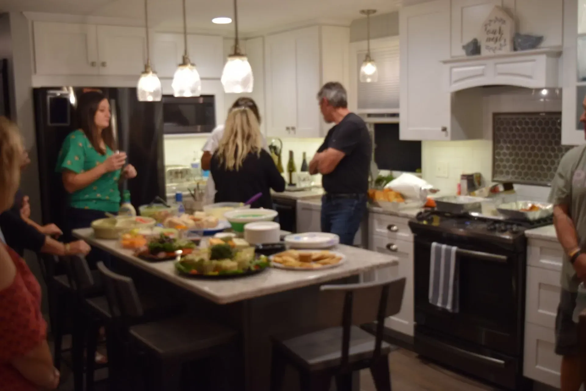 People gather in a bright white kitchen, food laid out on an island. Someone is near the stove.