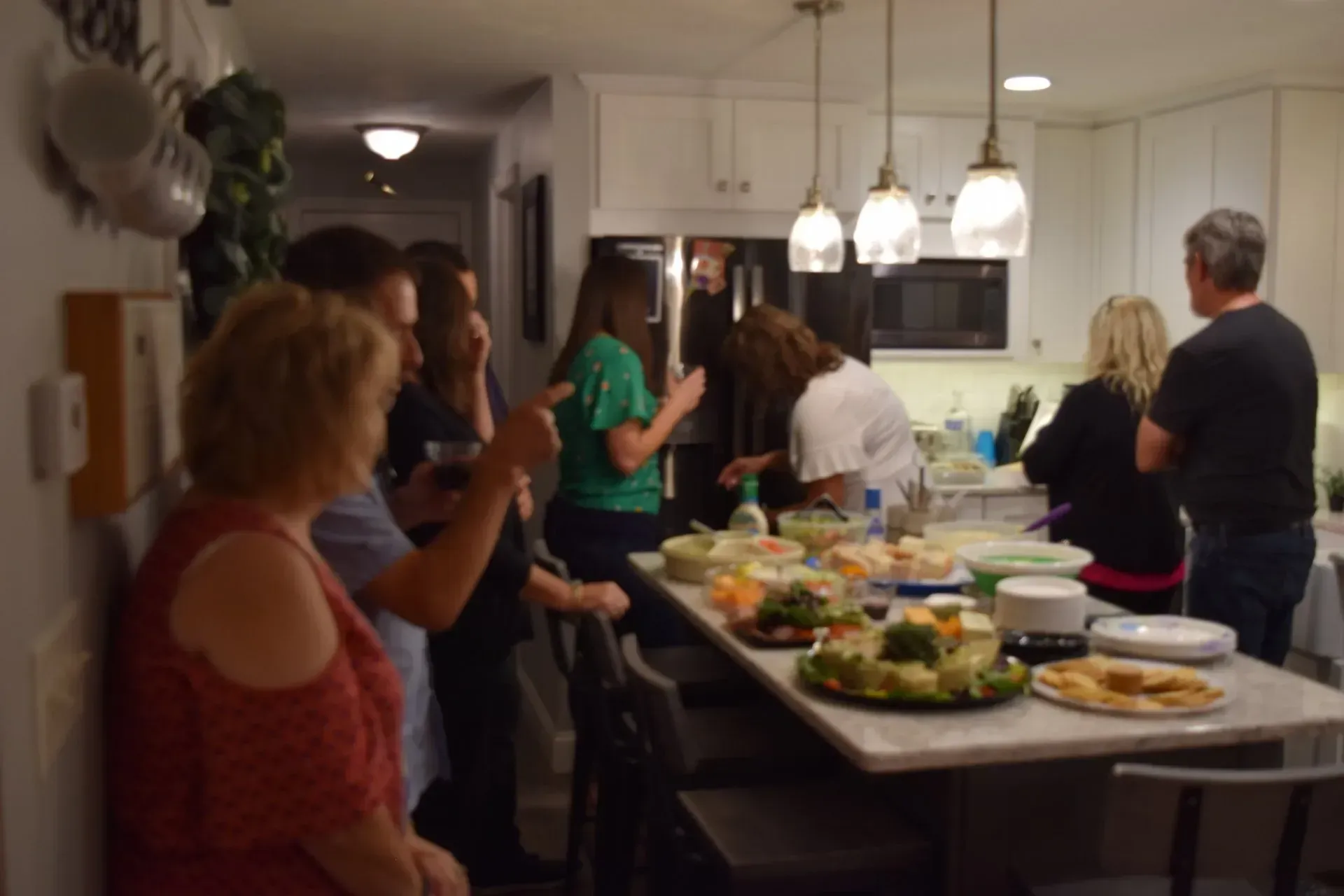 People gathered in a kitchen around a food-laden counter. Overhead lights illuminate the space.