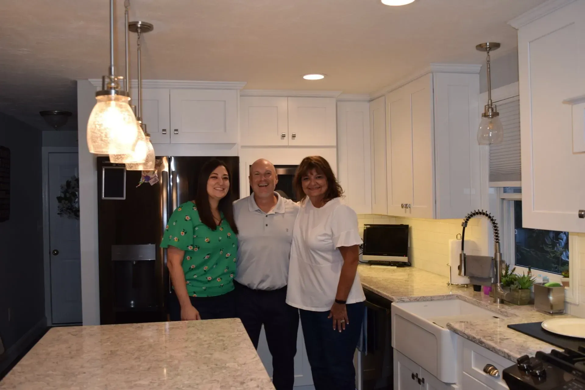 Three people smiling in a bright white kitchen with overhead lighting and granite countertops.