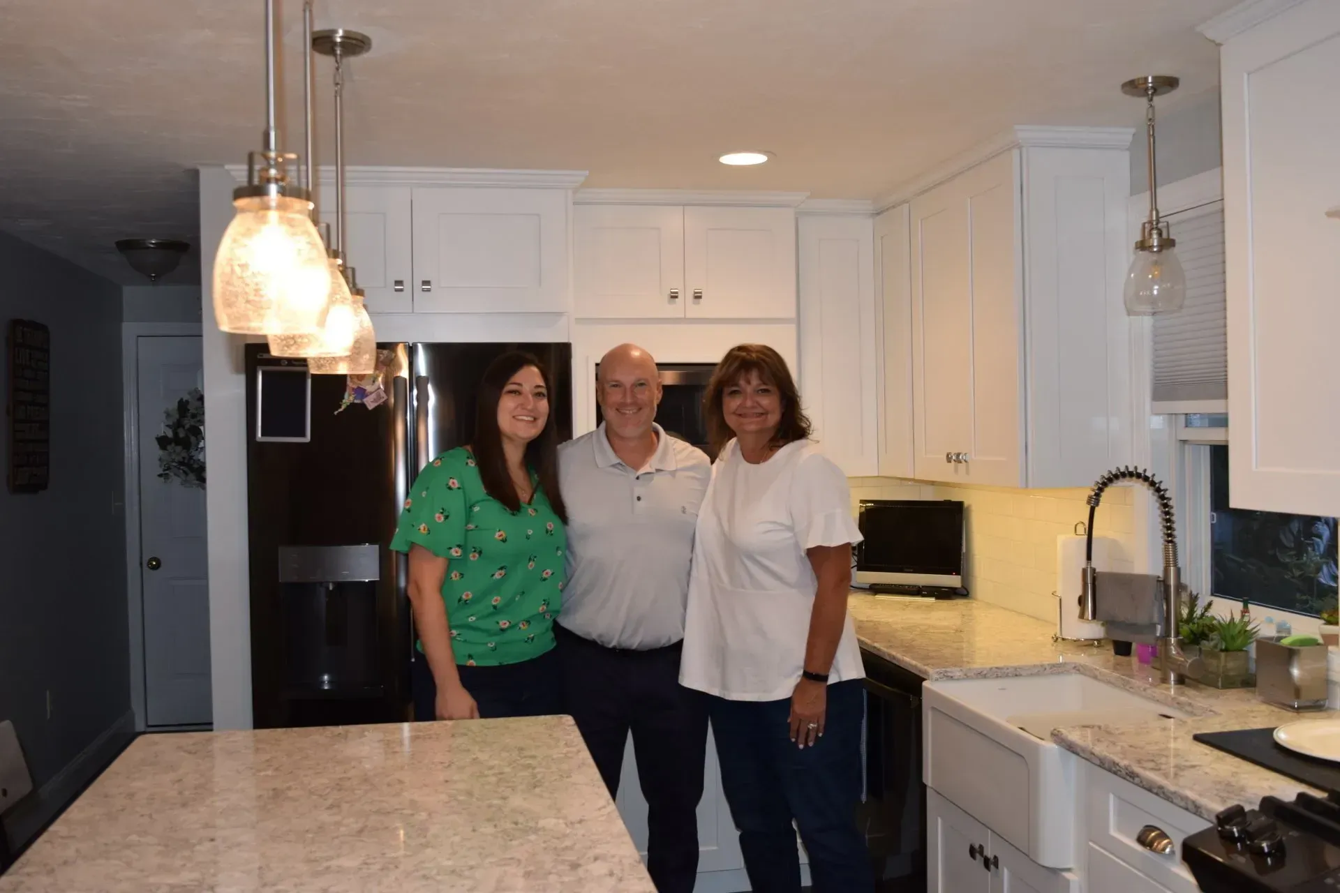 Three people smiling in a bright white kitchen with a large island and pendant lights.