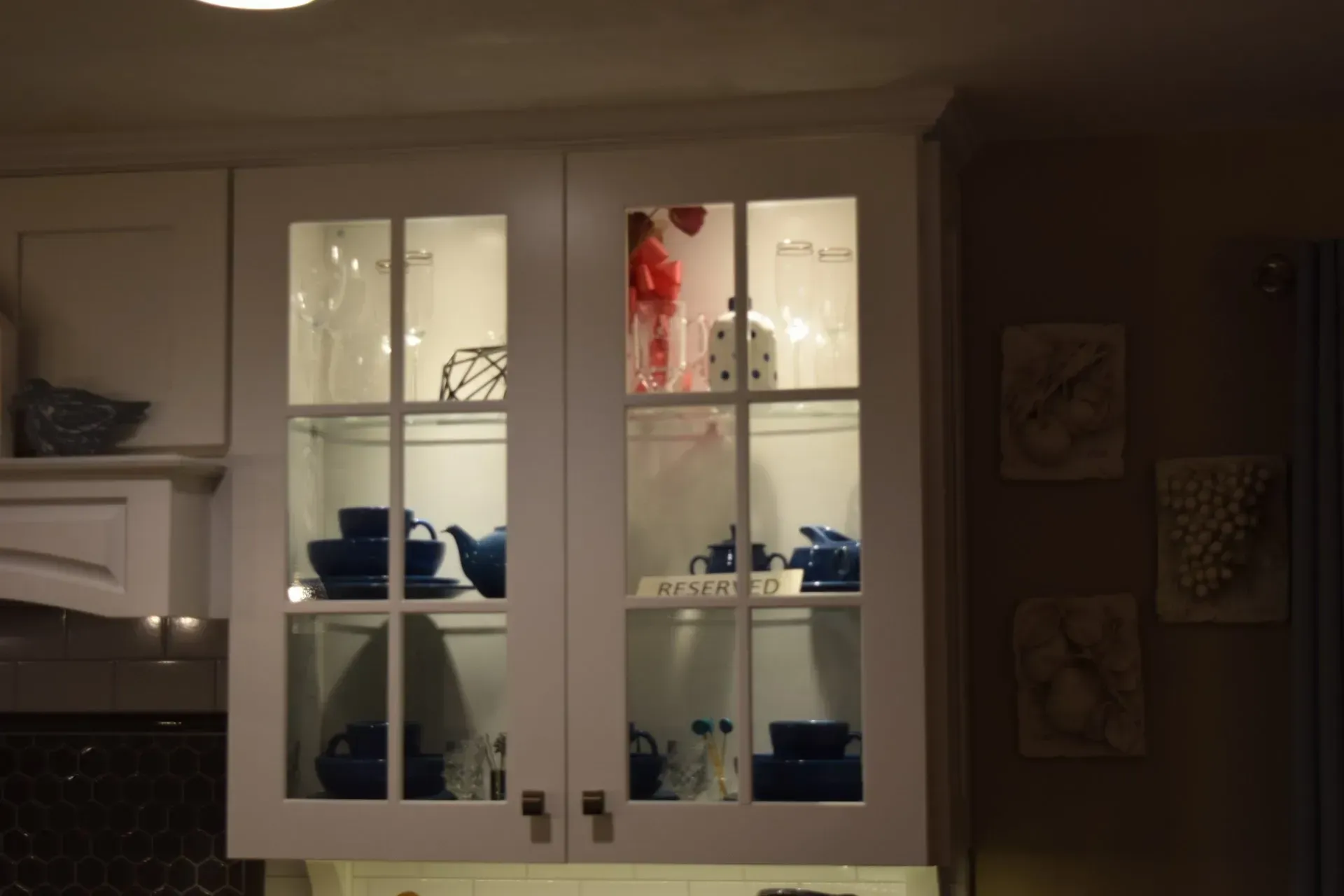 White kitchen cabinet with glass-paneled doors, lit from within, displaying blue dishware and decorative items.