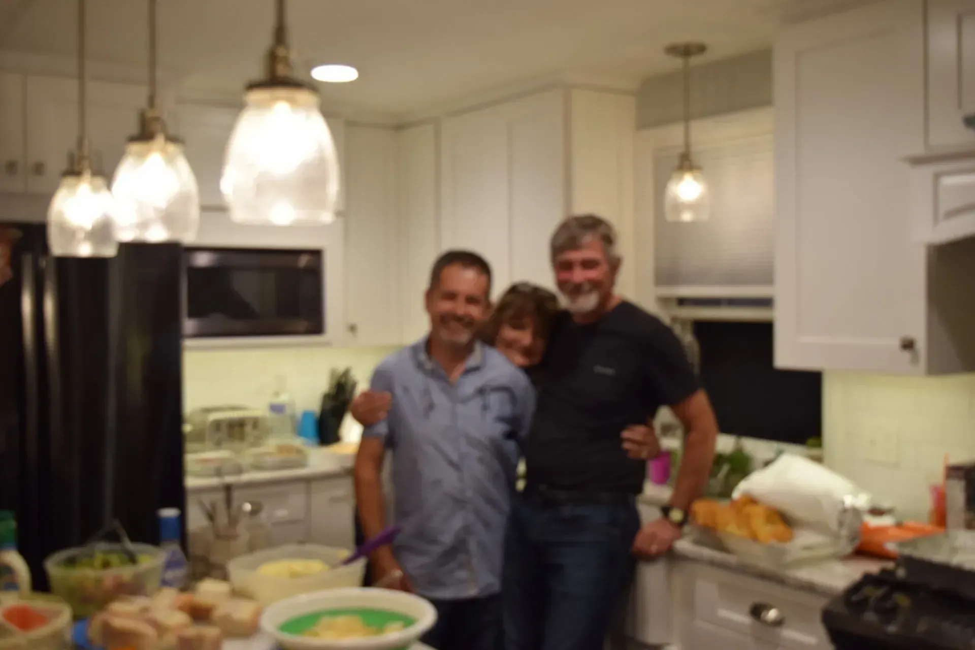 Three people smiling in a bright kitchen with white cabinets and pendant lights, food on the counter.