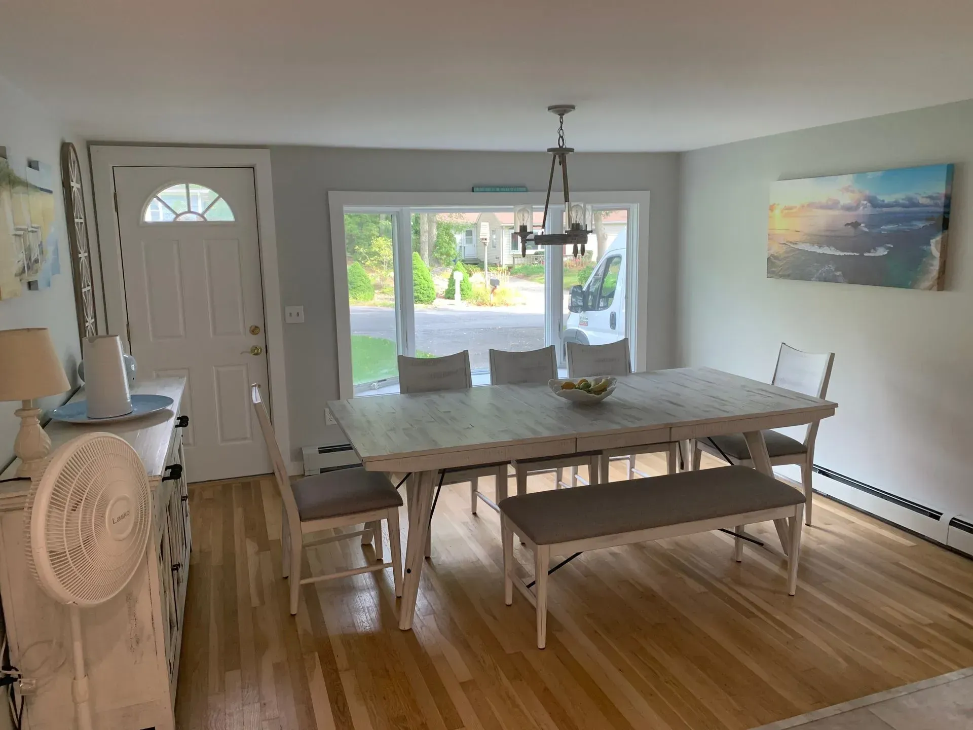 Dining room with wooden table and chairs, sunlight through a window, art on the wall.