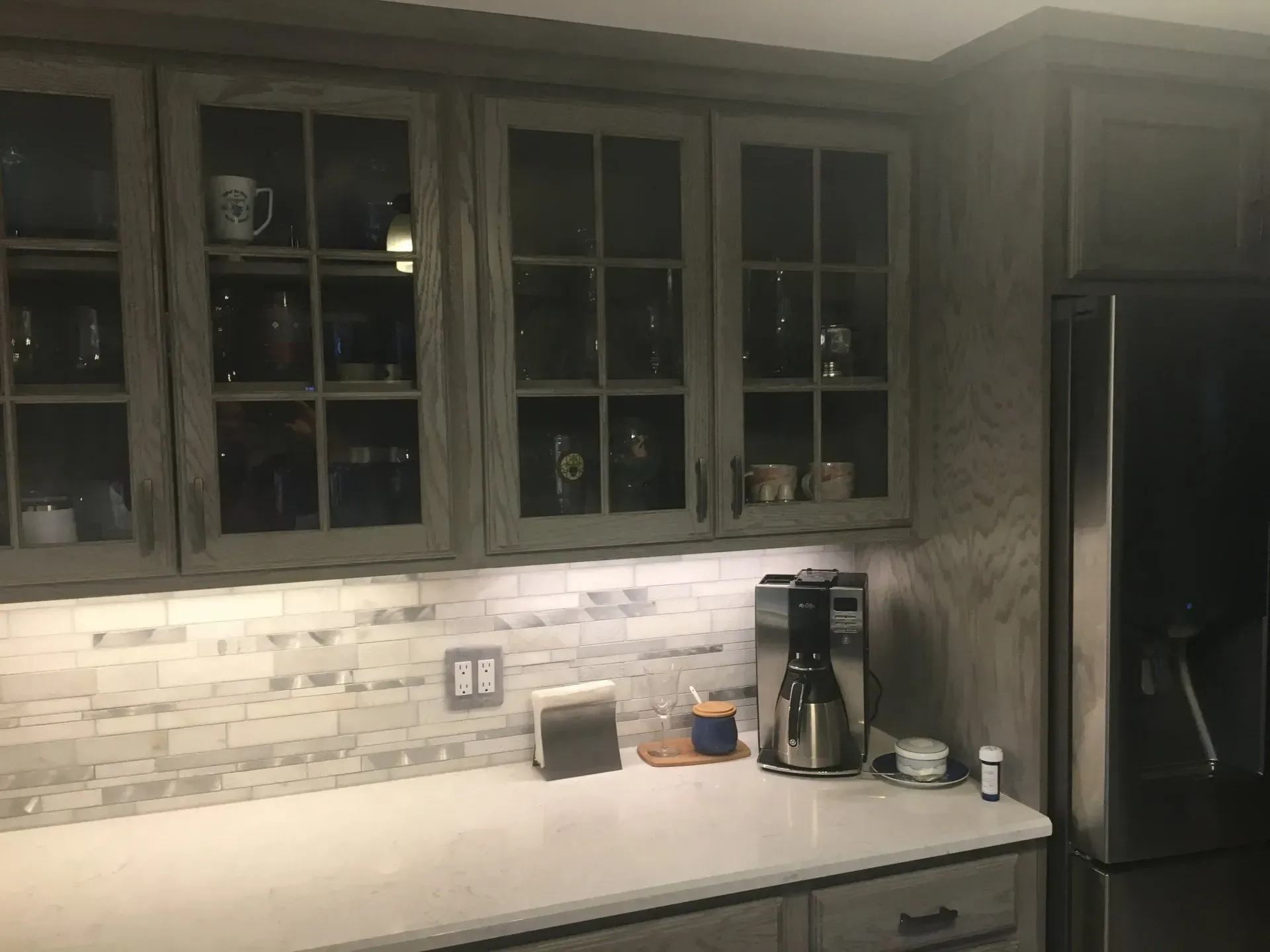 Kitchen cabinets with glass doors, white countertop, marble backsplash, coffee maker, and stainless steel refrigerator.