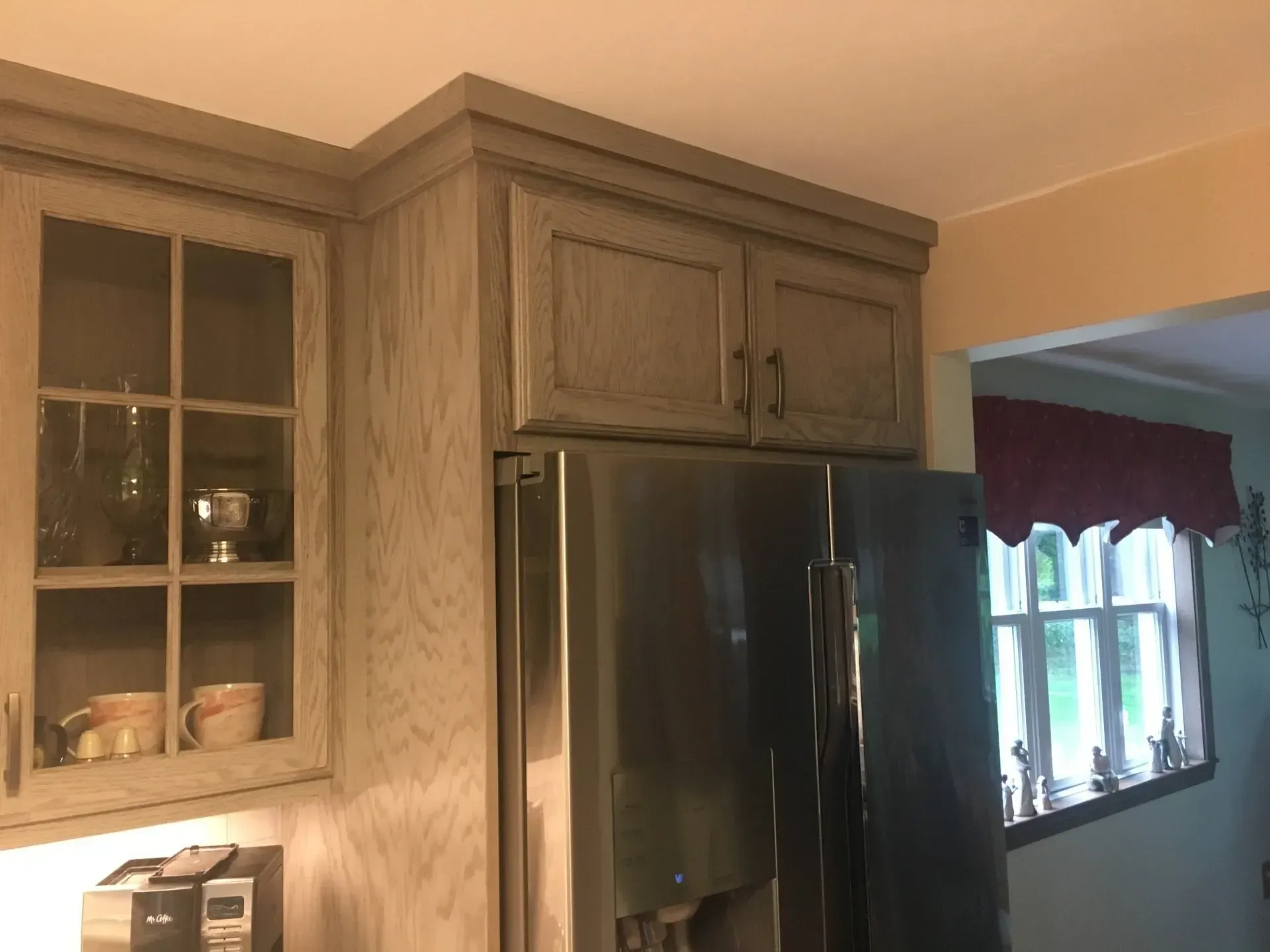 Kitchen cabinets, wood-tone, above stainless steel refrigerator. Corner cabinet with glass doors on the left.