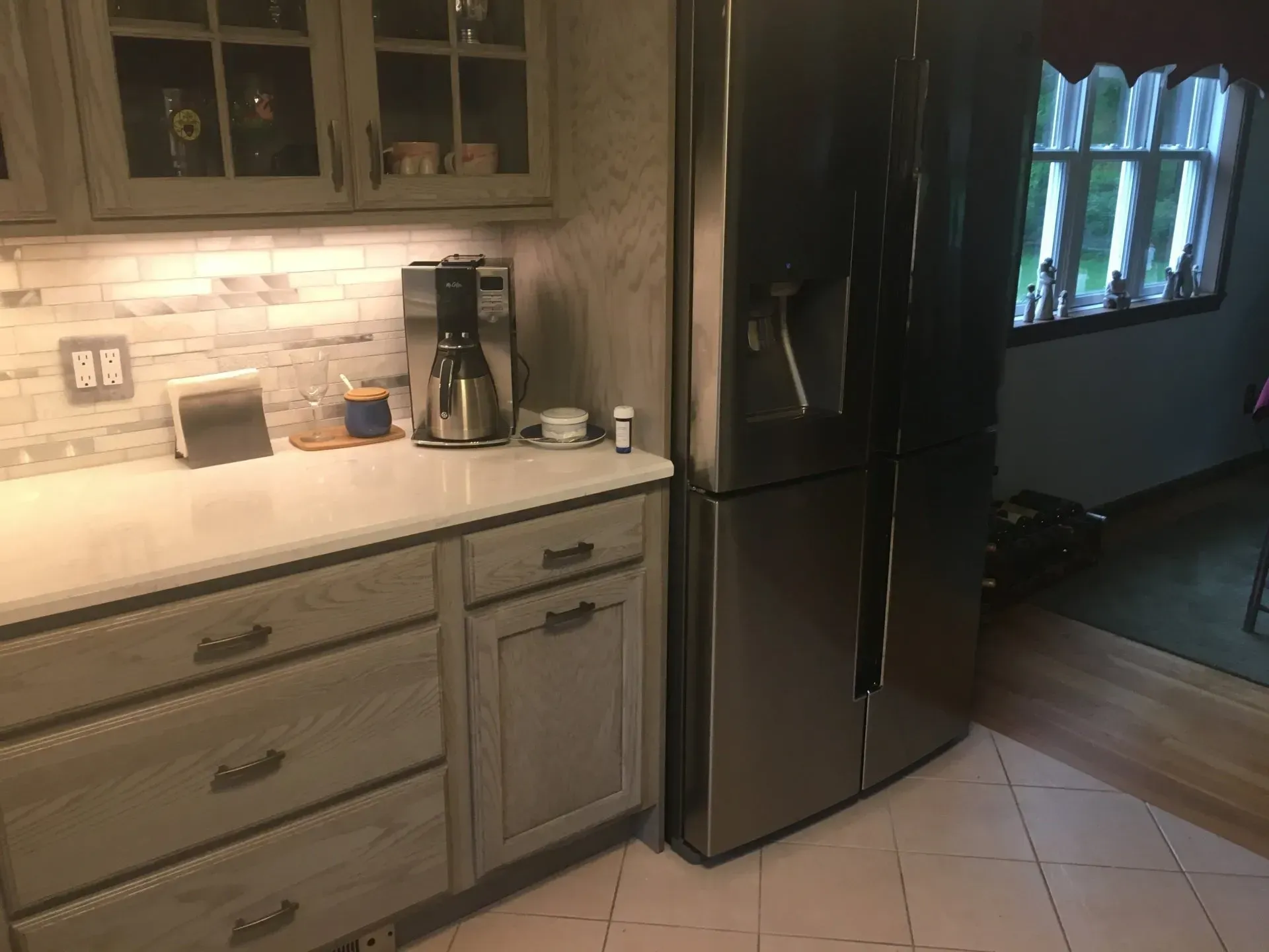 Kitchen with gray cabinets, white countertop, stainless steel refrigerator, and window.