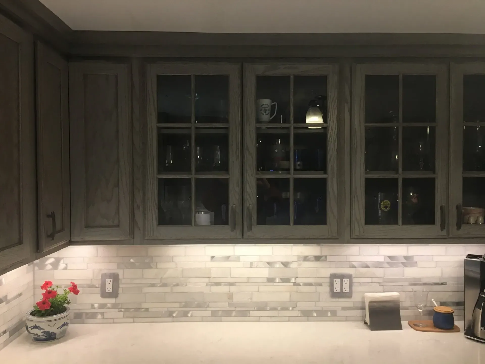 Kitchen cabinets, gray-brown with glass-fronted cabinets. White countertop with backsplash, under-cabinet lighting.
