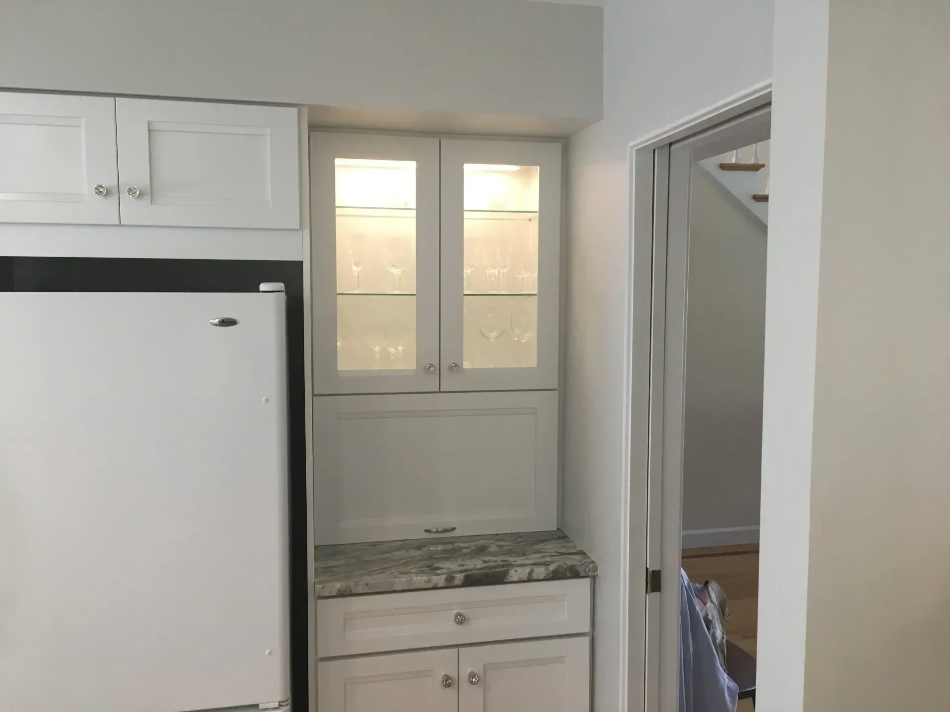 White kitchen cabinets and refrigerator. Glass-front cabinet over granite countertop. Doorway on right.
