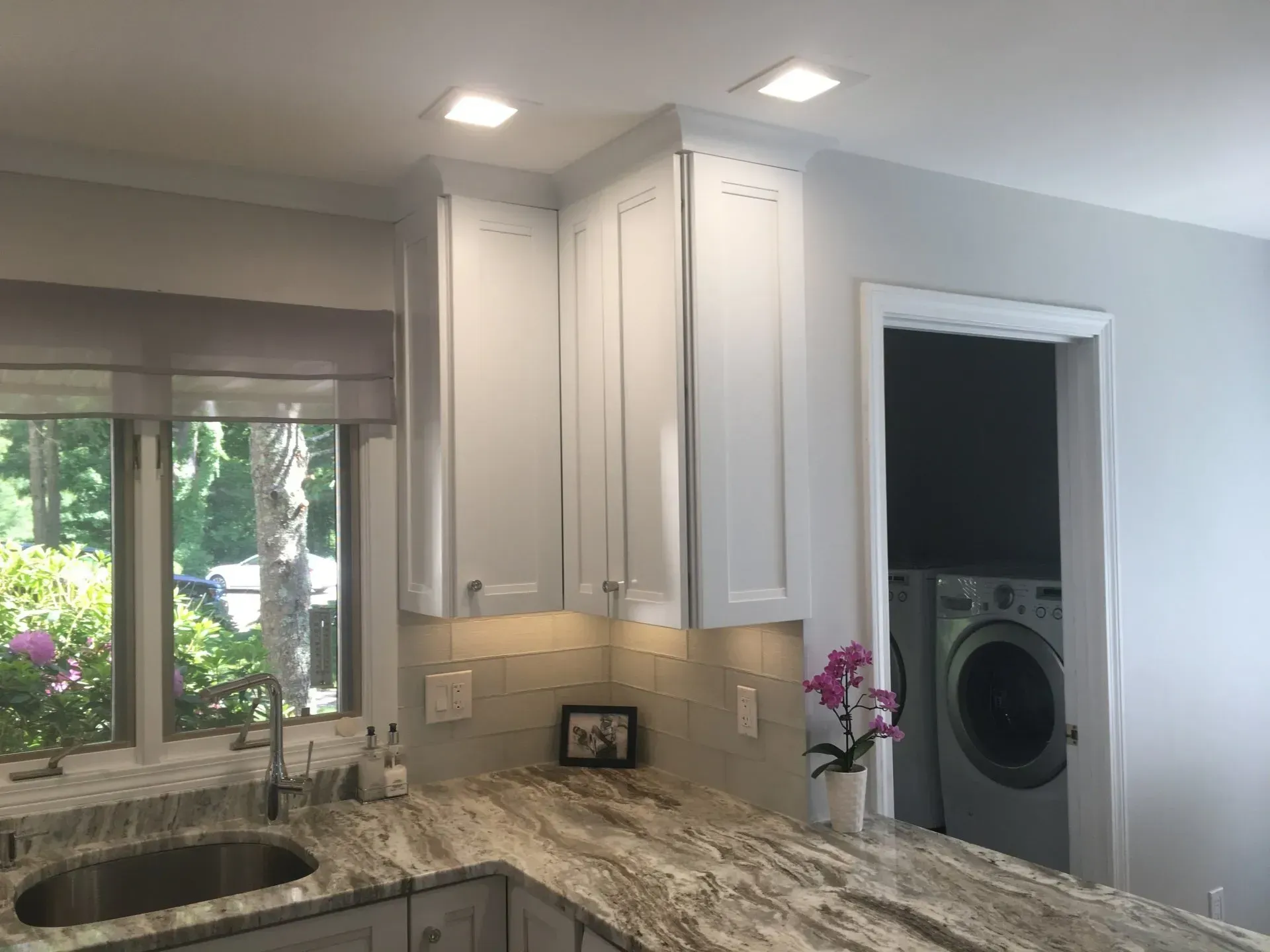 Kitchen corner with white cabinets, granite countertop, sink, window, and a washing machine visible in doorway.