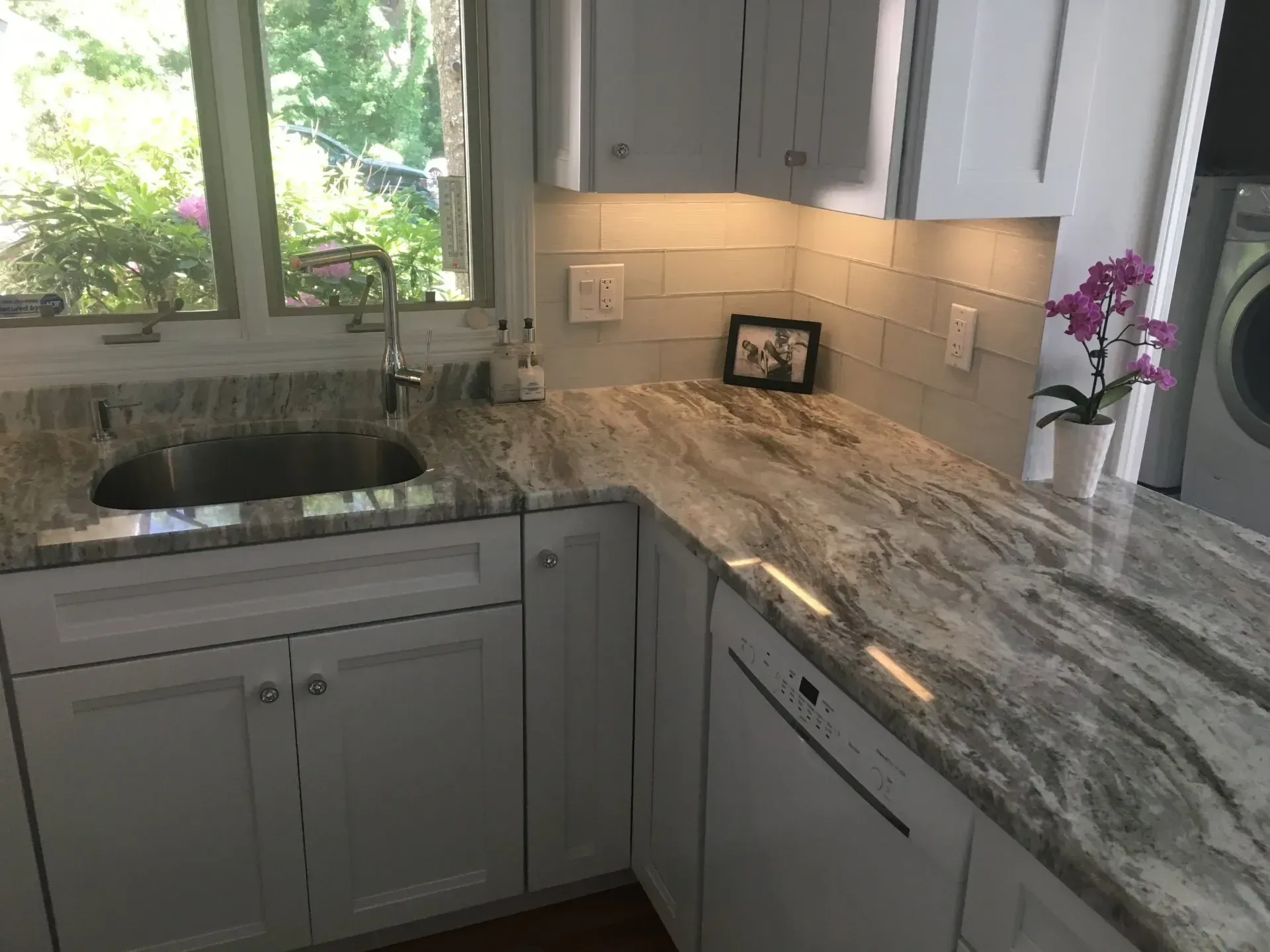 White kitchen cabinets with granite countertop, stainless sink, and dishwasher. Bright natural light.