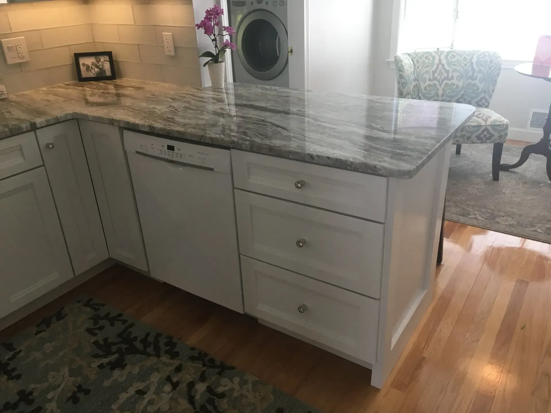 White kitchen island with granite countertop, dishwasher, drawers. Light wood floor, rug.