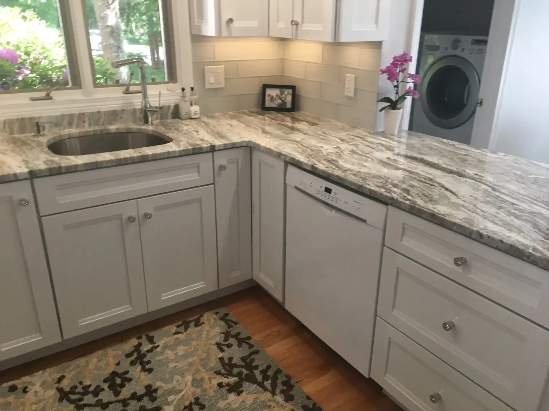 White kitchen with granite countertops, sink, dishwasher, cabinets, and window.