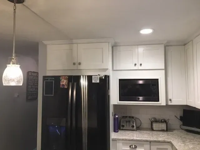 White kitchen cabinets above a black refrigerator and microwave. A pendant light hangs.