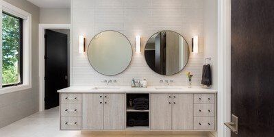 Bathroom with two round mirrors, sconces, light wood vanity, and black door.