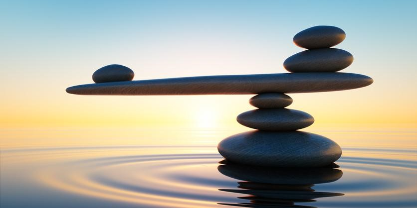 Stones balanced on each other, mimicking a scale, set against a calming water and sunset backdrop.