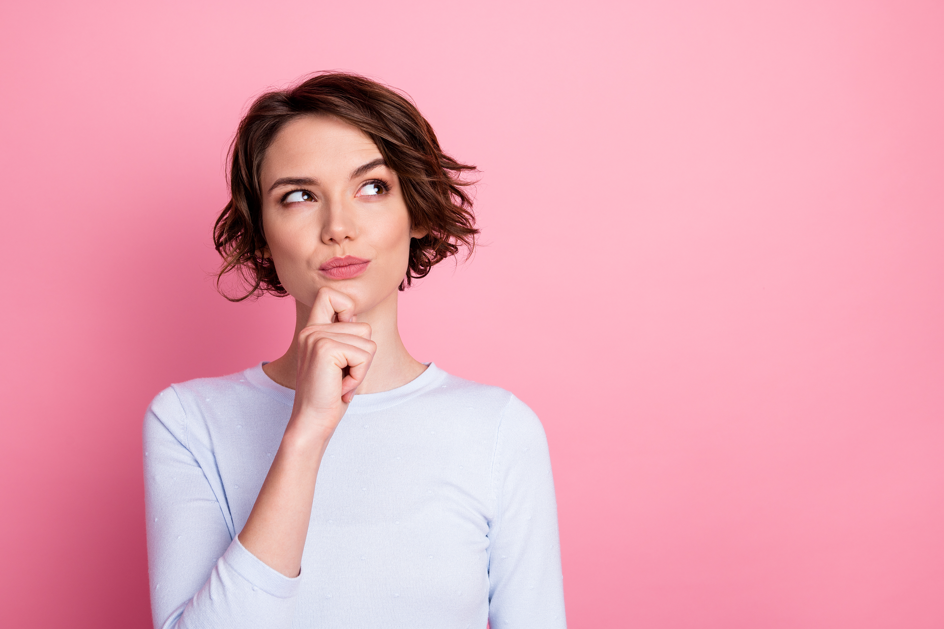 Woman with curly brown hair looking up thoughtfully, hand on chin, against a pink background.