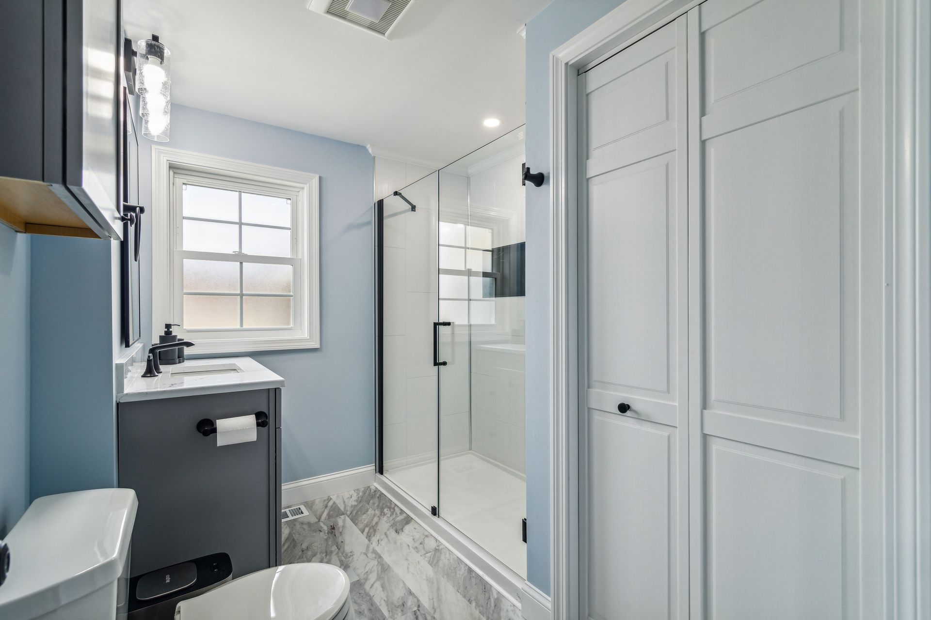 Blue bathroom with a shower, toilet, and vanity. White closet door and window.