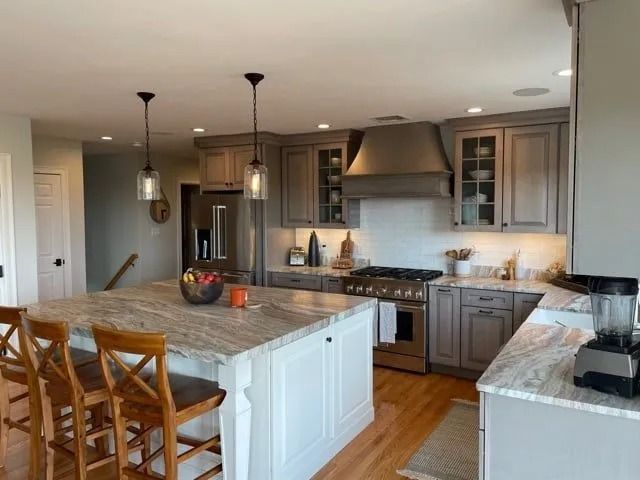 Modern kitchen with gray cabinets, marble countertops, stainless steel appliances, and a central island with bar stools.