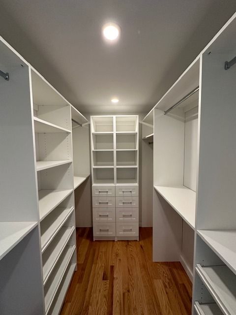 White custom walk-in closet with shelves, drawers, and hanging rods. Hardwood floor. Lit by overhead light.