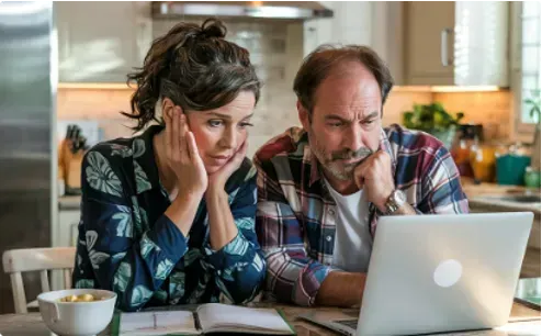 A couple, looking concerned, reviews information on a laptop in a kitchen.