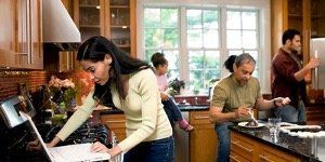 Woman on laptop in kitchen, others eating and interacting. Brightly lit with cabinets and a window.