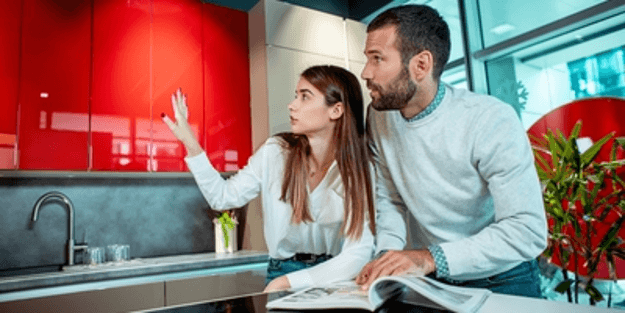A couple examines kitchen cabinets in a showroom. Woman gestures, man looks on. Red cabinets visible.