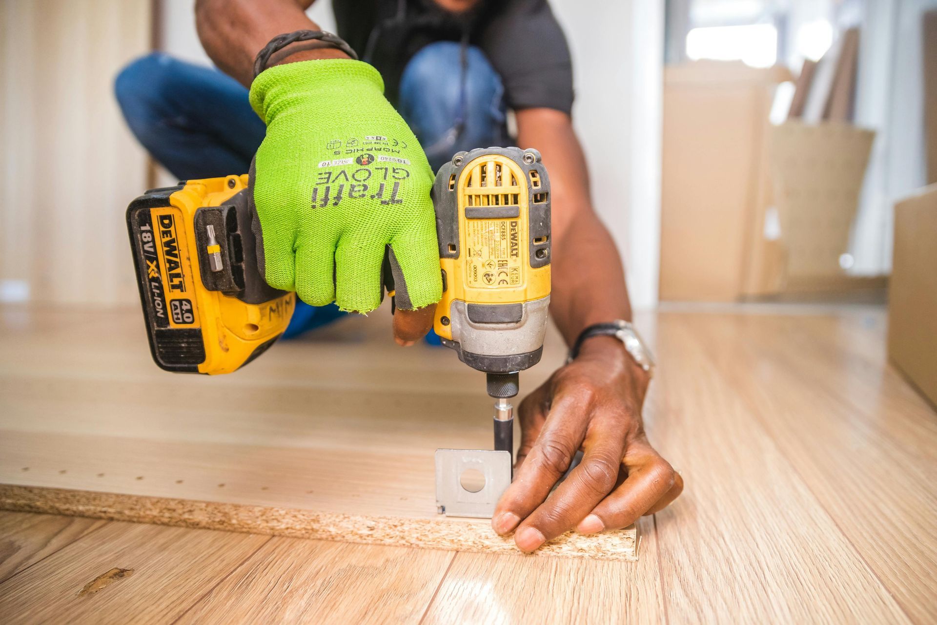 Person using a power drill on a wooden surface, wearing a green work glove.