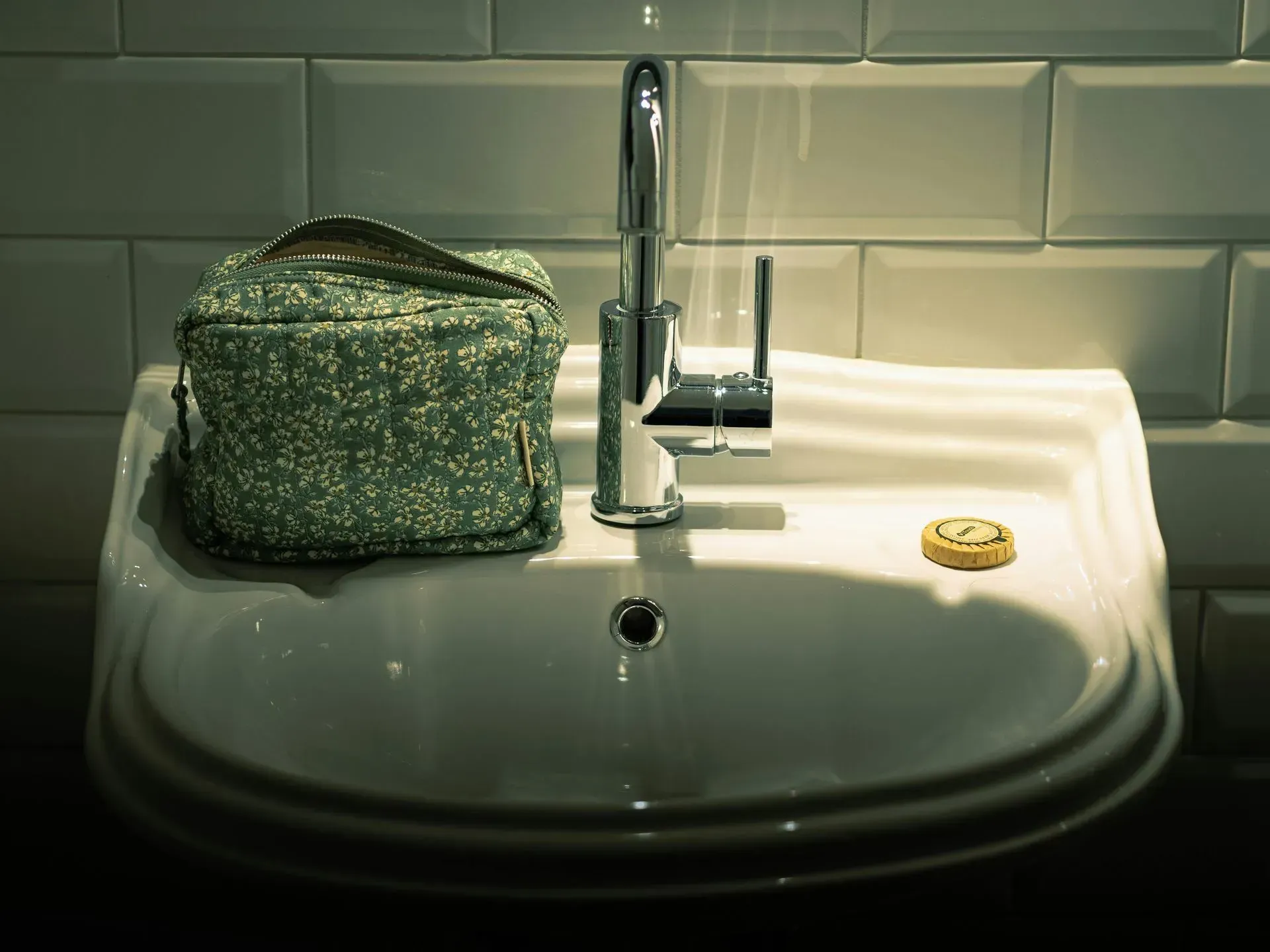 White sink with silver faucet. A green cosmetic bag sits on the left. A small round object is on the right.