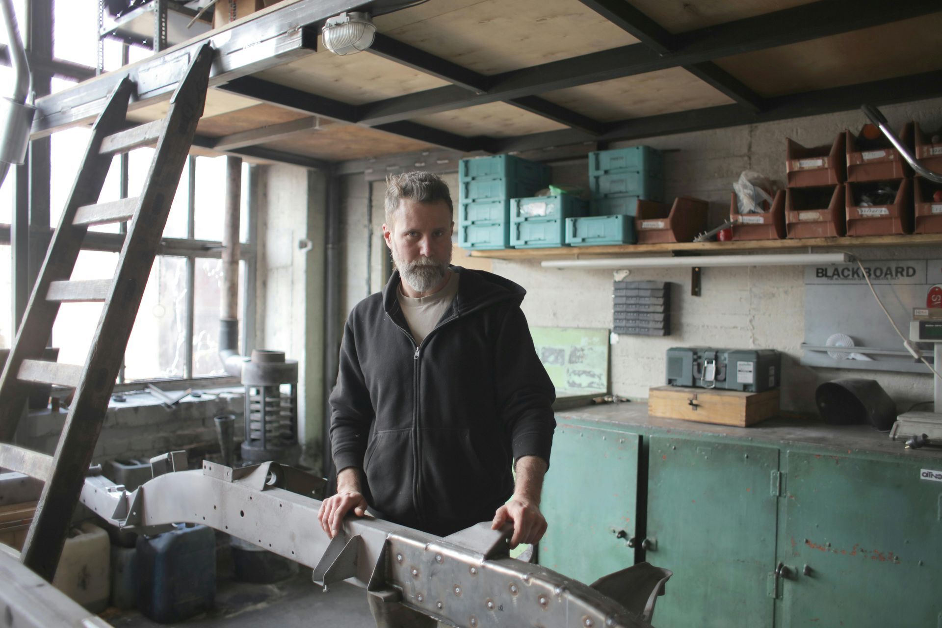 Man in workshop holding metal frame; shelves with bins in background.