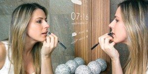 Woman applying makeup in front of a mirror with digital time display.