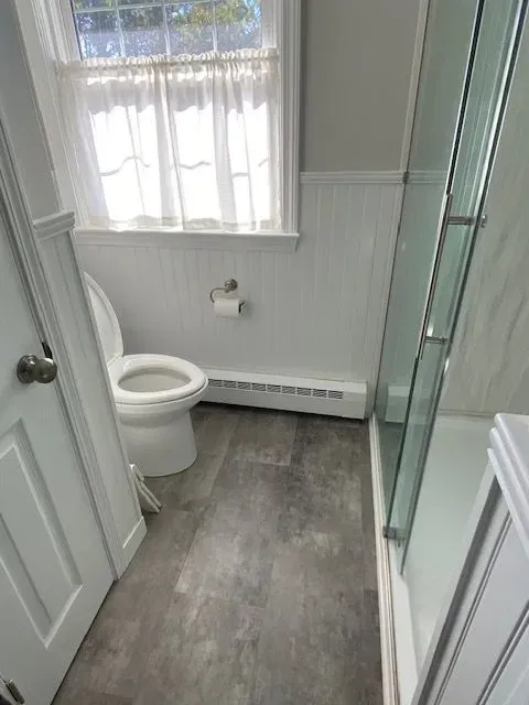Bathroom interior with toilet, shower, window, and gray floor. White paneling and door.