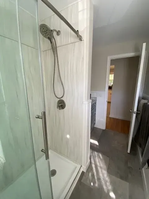 Shower with glass door, handheld showerhead, light gray walls, open doorway to hallway.
