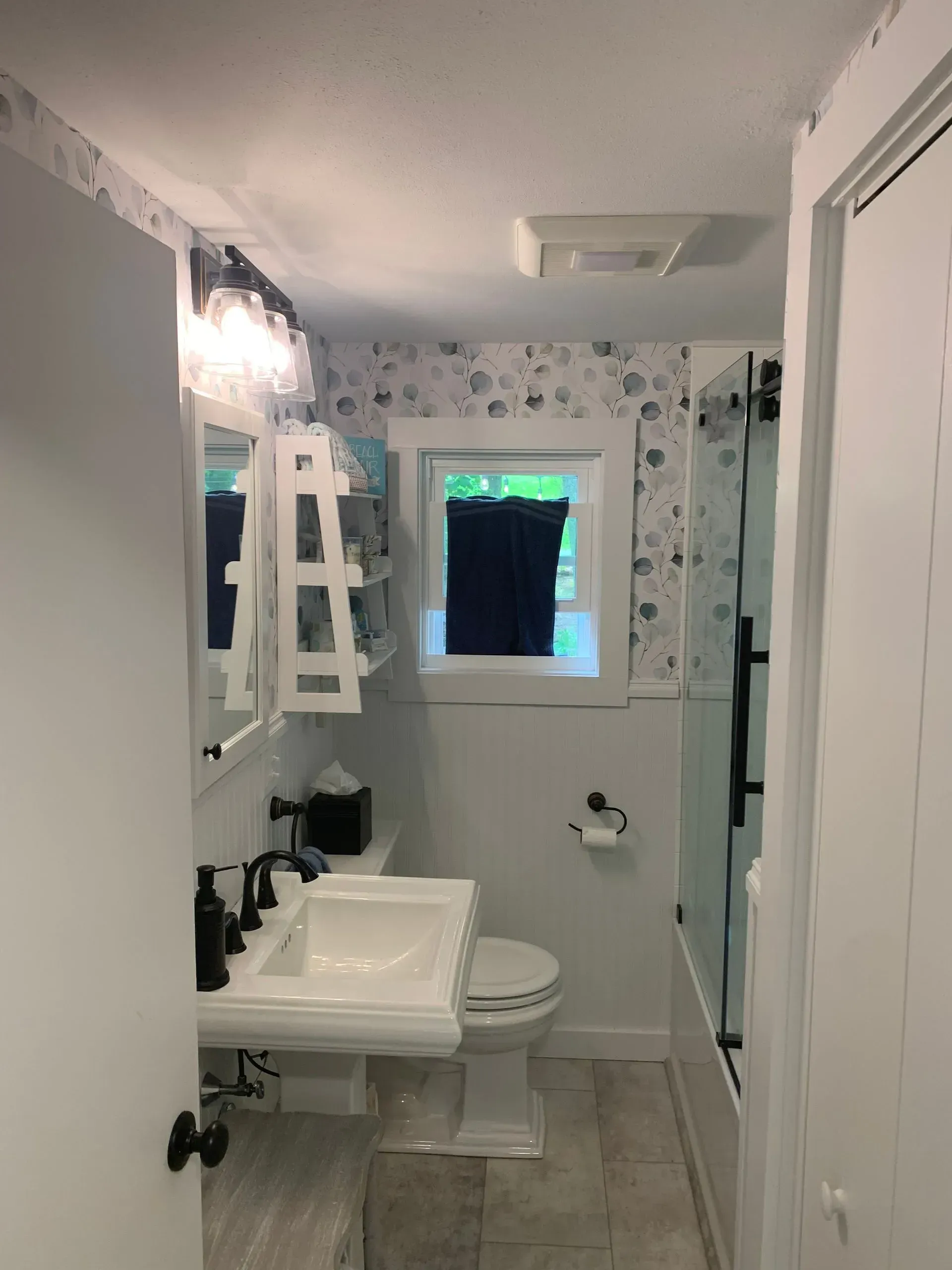 A small white bathroom with a pedestal sink, toilet, and shower. Shell patterned wallpaper accents.