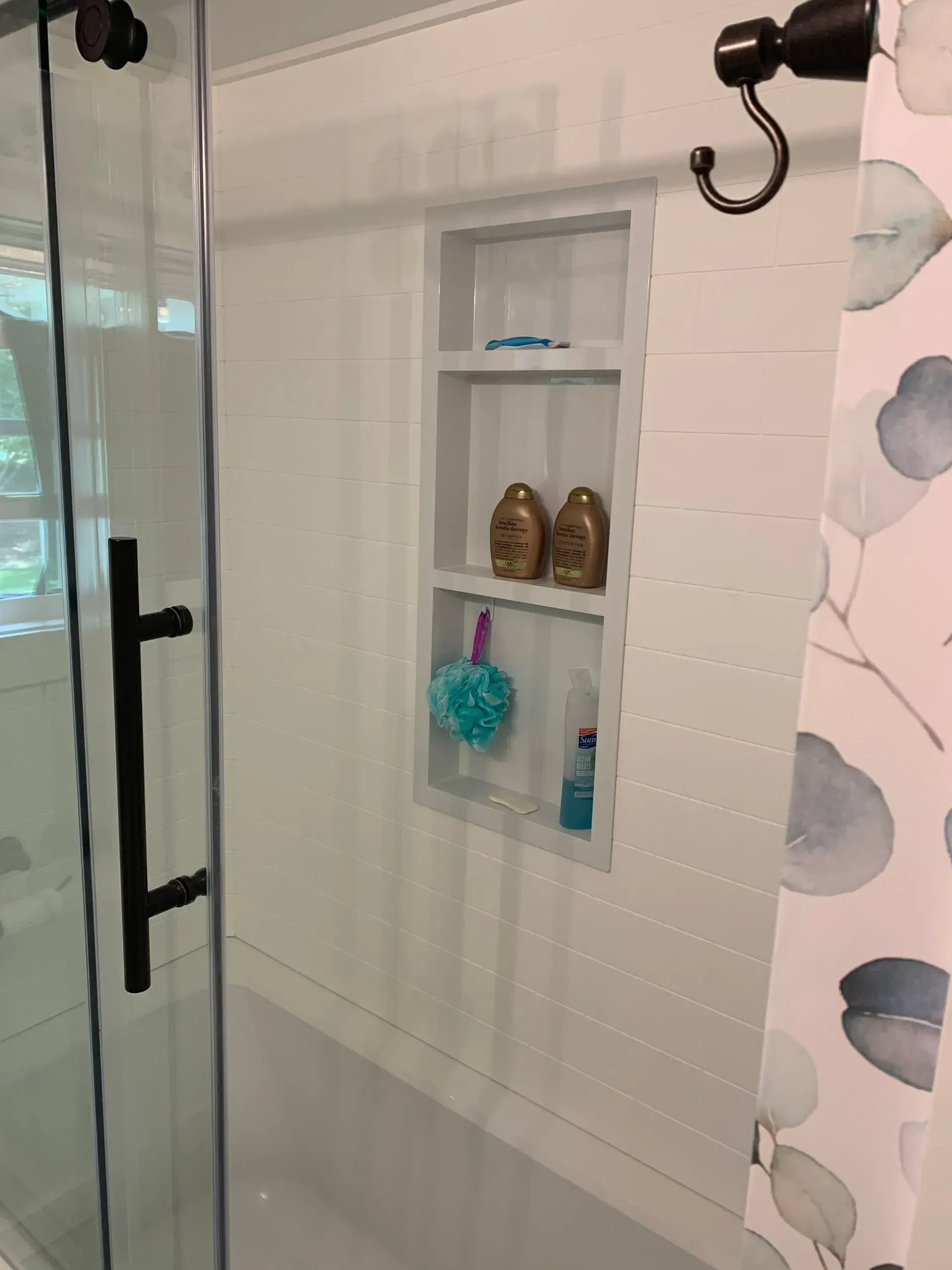 White shower with built-in shelf holding soap, loofah, and shampoo. Glass door and shower curtain are present.