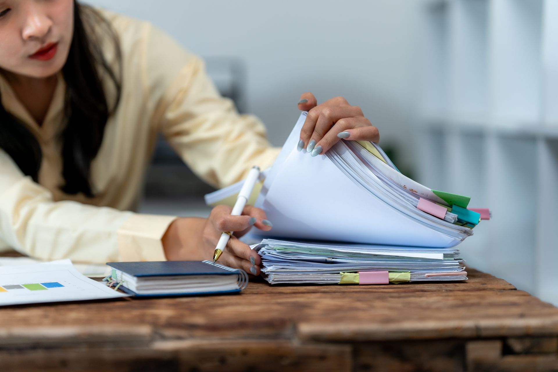 Woman at a desk sorting through a stack of papers with a pen, notebook, and chart nearby.