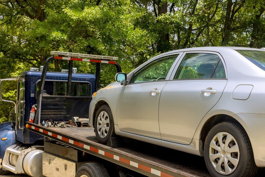 Silver sedan on a tow truck with a blue car behind it, parked under trees