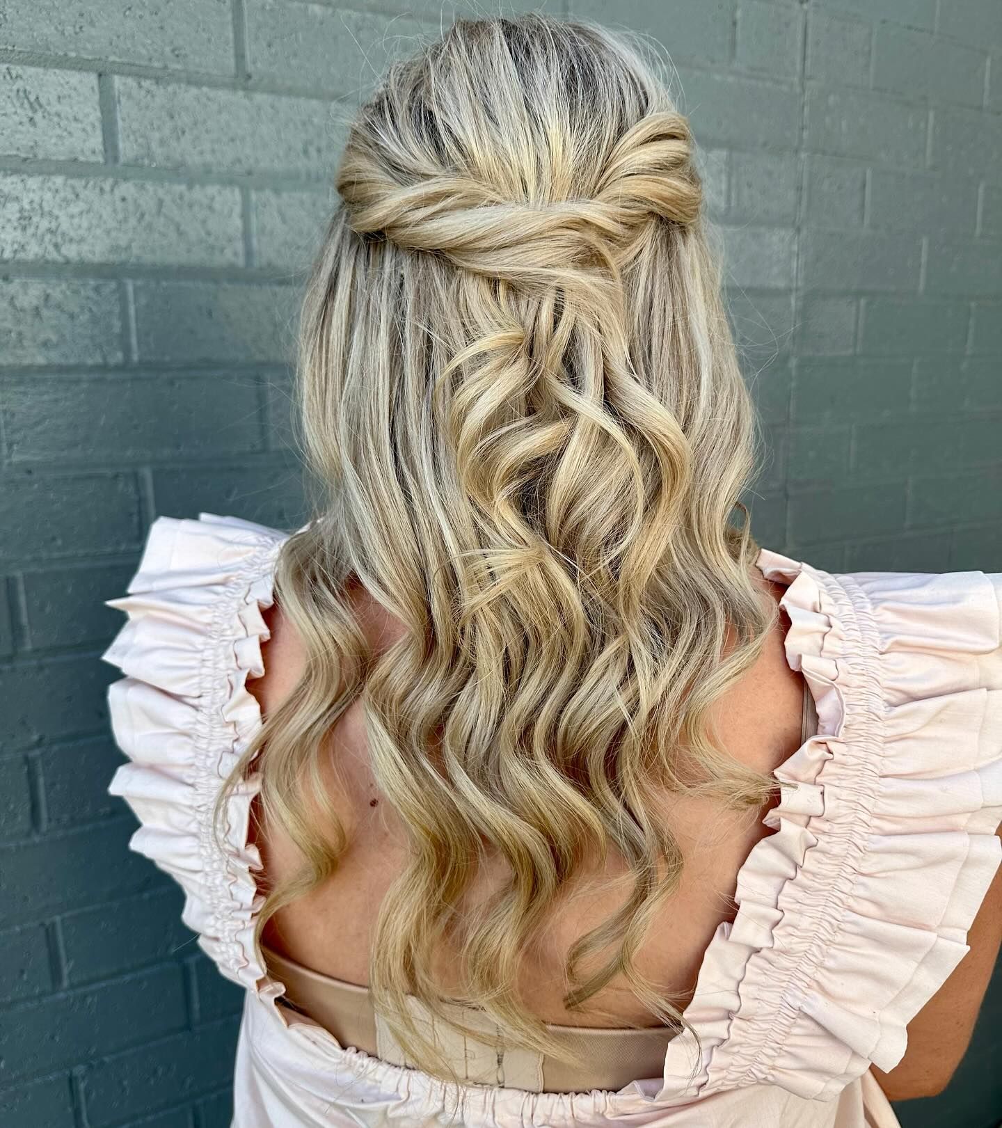 Long blonde wavy half-up hairstyle with a braided crown and loose curls against a teal brick wall — Pose Hair Design in Orange, NSW