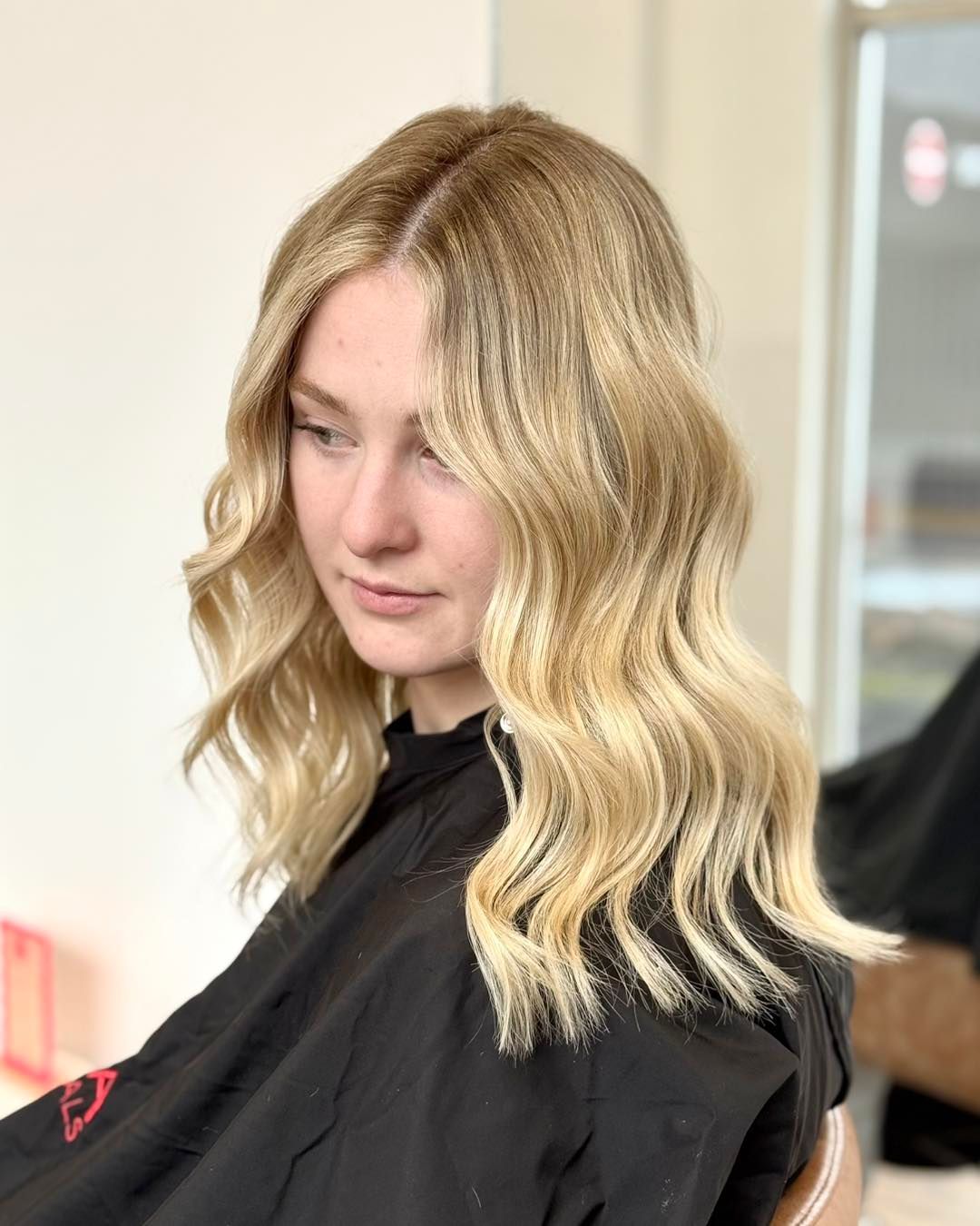 Woman with wavy blonde hair in a salon chair, wearing a black cape, looking down — Pose Hair Design in Orange, NSW