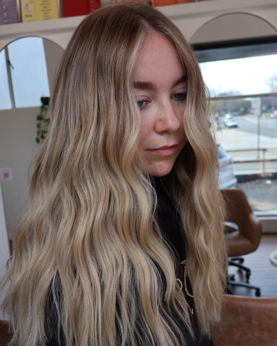 Woman with long wavy blonde hair in a salon, looking down beside a mirror — Pose Hair Design in Orange, NSW