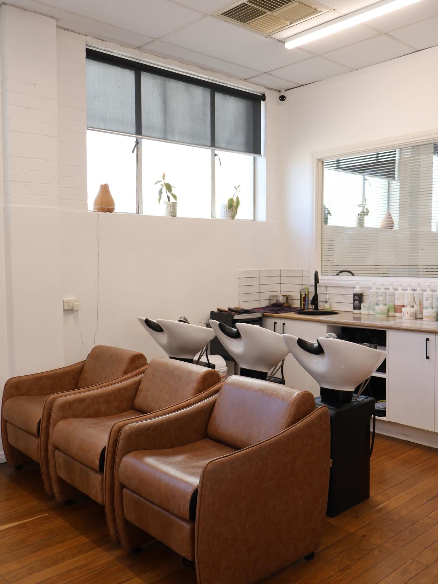 Salon waiting area with two brown chairs, shampoo sinks, mirrors, and a window with blinds — Pose Hair Design in Orange, NSW