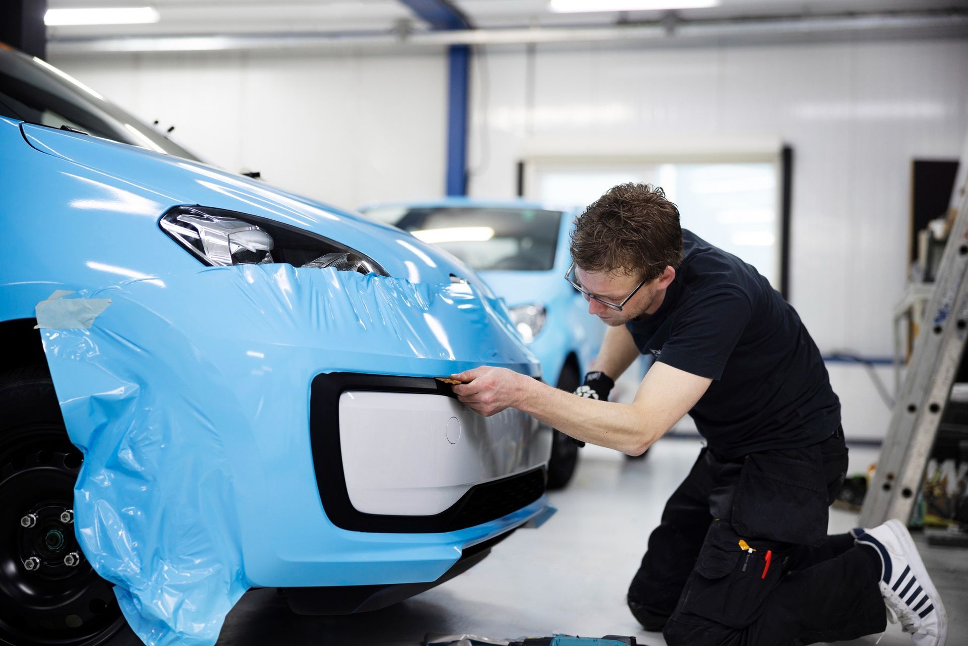 A man is wrapping the front of a blue car with plastic wrap.