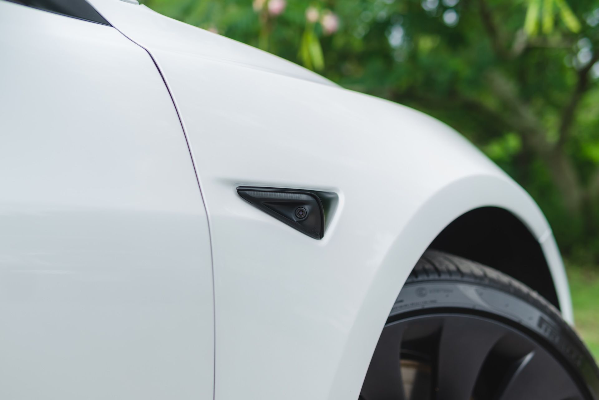 White Tesla car's front fender detail with black side marker and wheel; green foliage in background.
