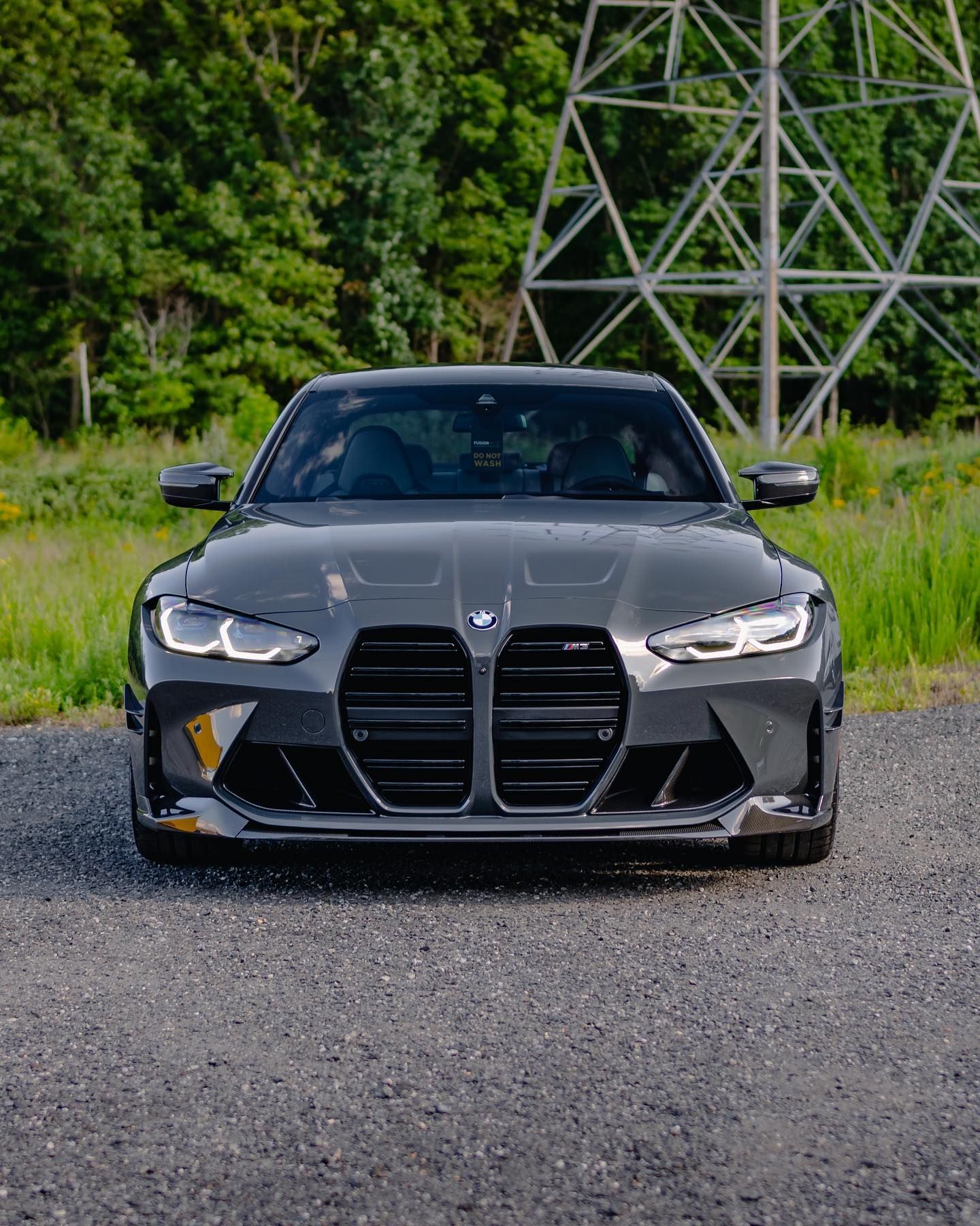 Gray BMW M4 sports car parked on gravel, facing forward. Trees and a metal structure are in the background.