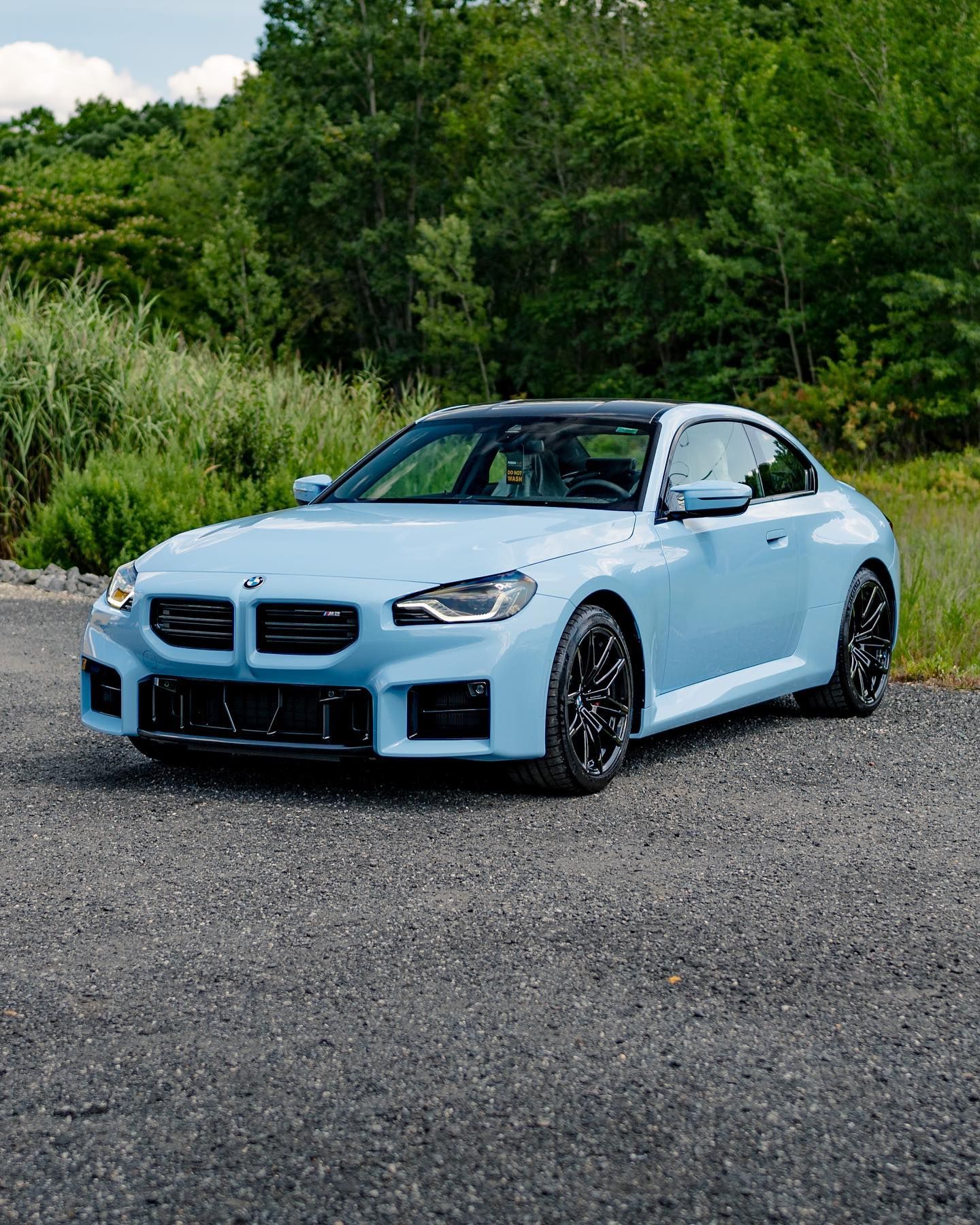 Light blue BMW M2 coupe parked on gravel with a forest background. Black wheels.