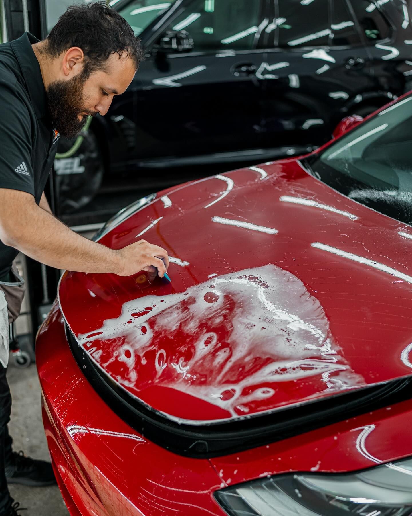 Man applying film to red car hood in a shop, using a squeegee and soapy solution.
