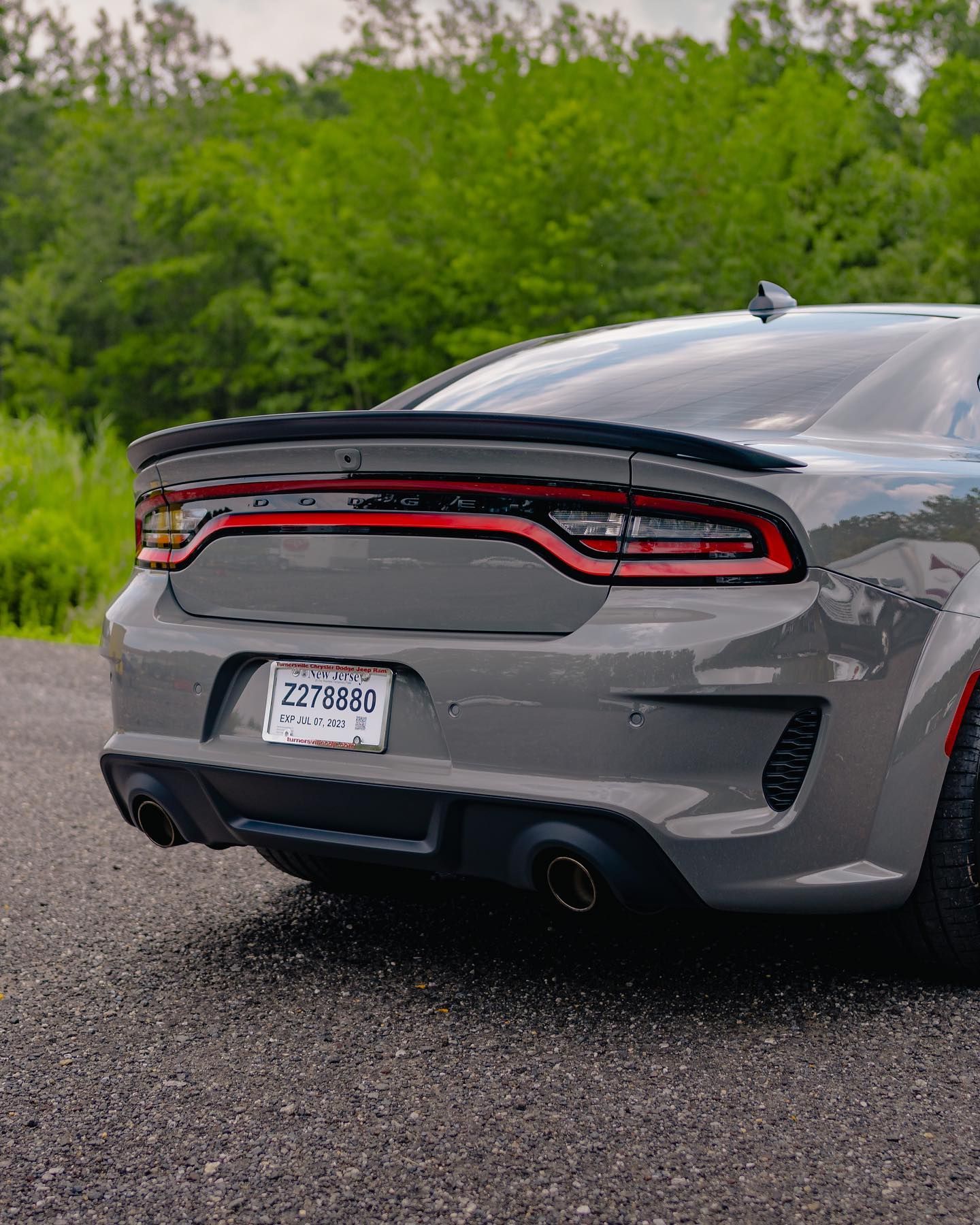Gray Dodge Charger rear view, black spoiler, dual exhaust, license plate, gravel road, green trees.