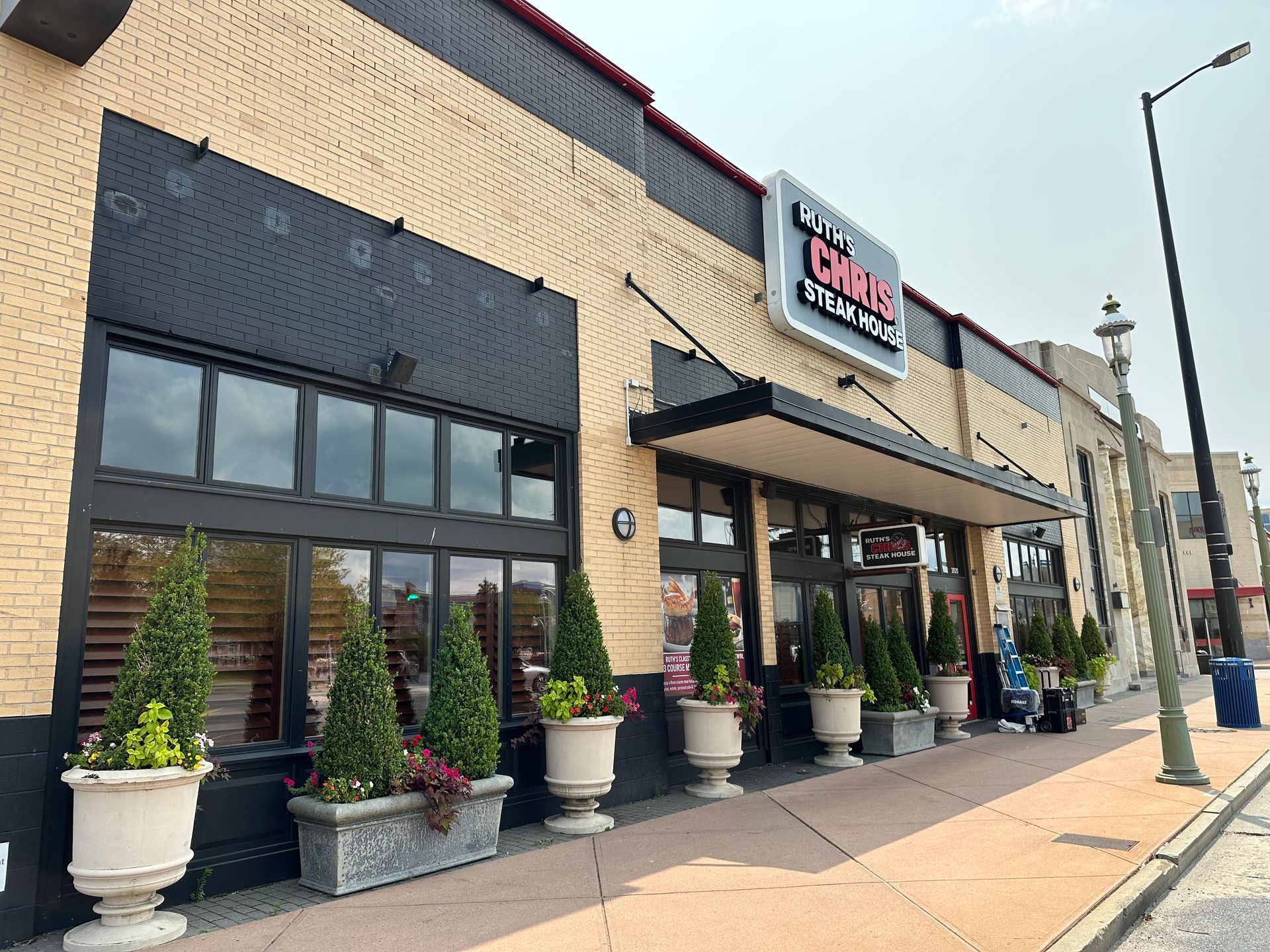 Restaurant facade with black trim, tan brick, sign "City Barbeque," potted plants, and street.